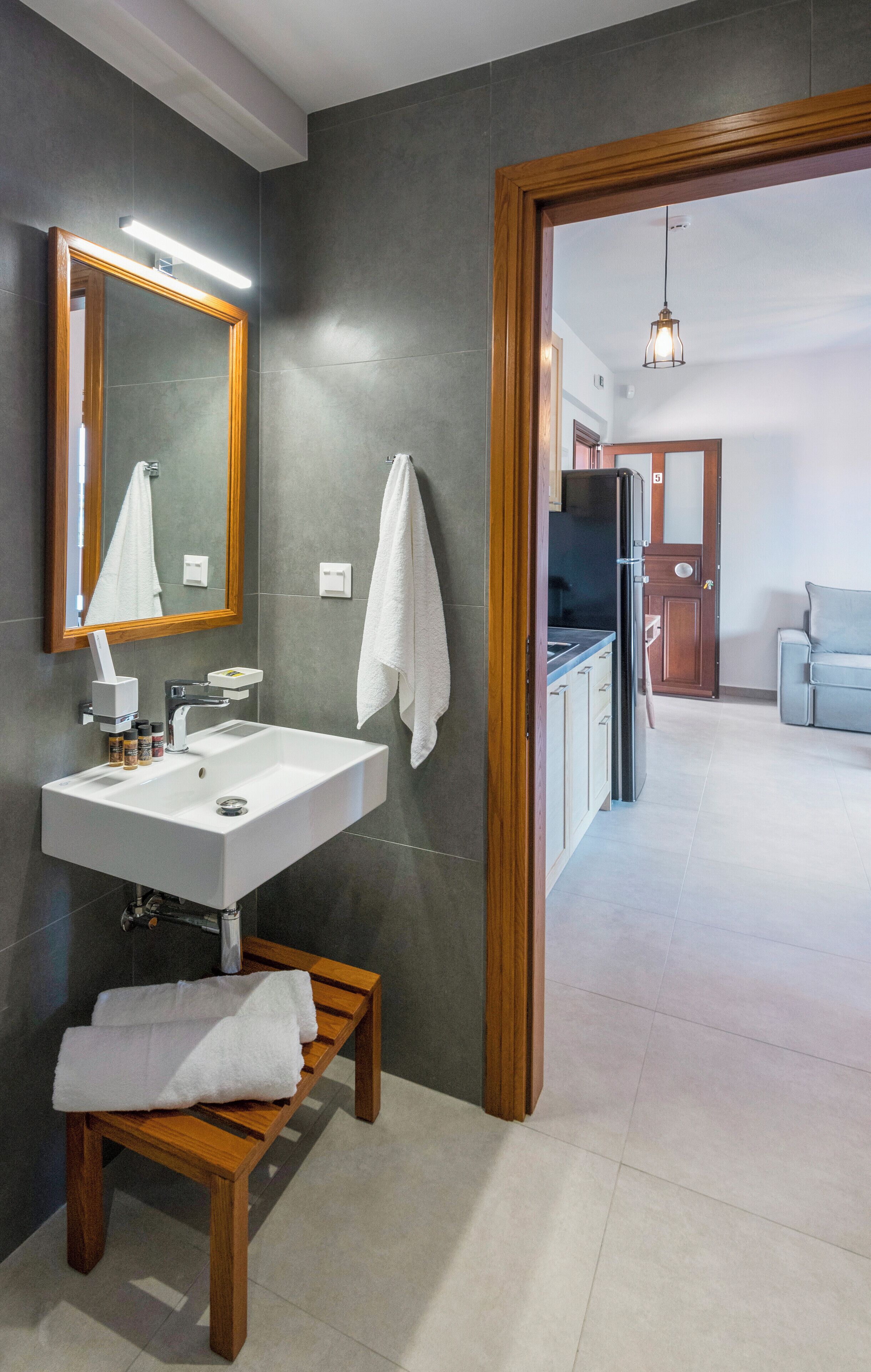 A modern bathroom with a white sink, wooden bench, and a large mirror.