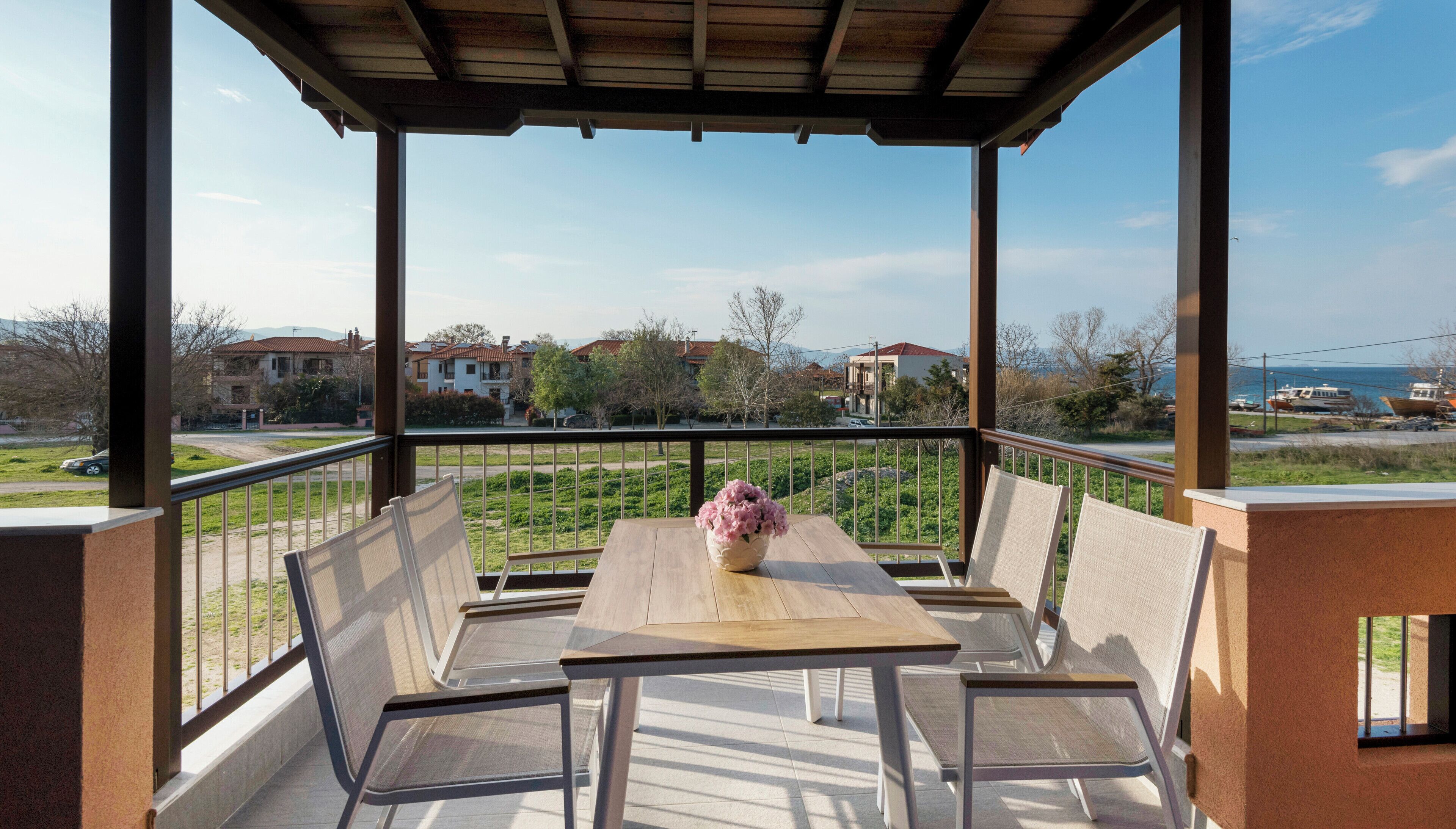 Un balcón techado con una mesa y sillas, un florero con flores, y vistas a casas y barcos a lo lejos.