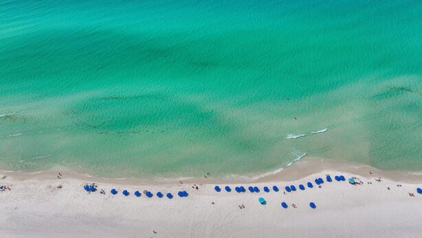 Luchtbeeld van een strand met helder turquoise water en een zandstrandrand met blauwe parasols.