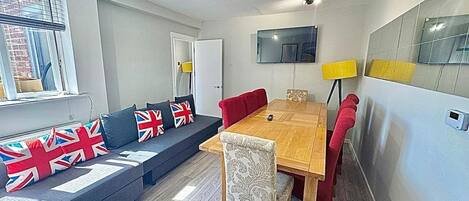 Een moderne woonkamer met een zithoek, een eettafel en een aan de muur bevestigde televisie.