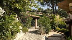 En sti af brosten fører til en traditionel bygning med tegltag, omgivet af frodig vegetation og blomstrende planter.