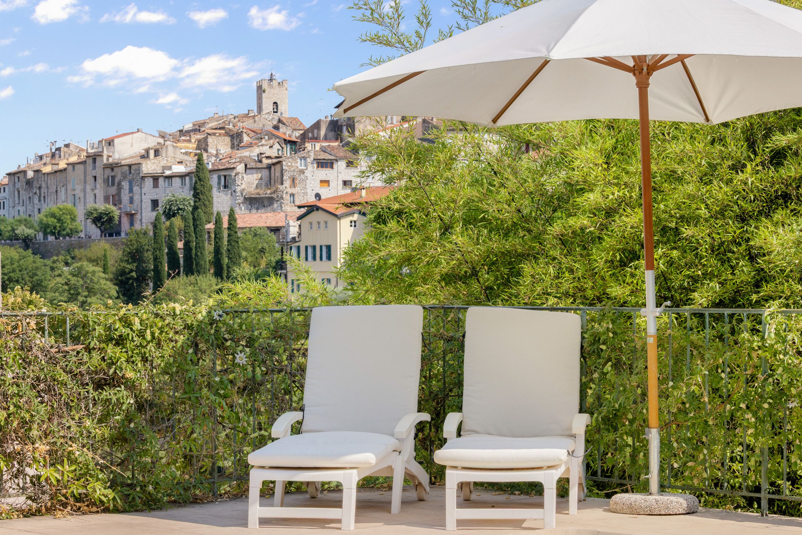 Deux fauteuils de jardin blancs, accompagnés d’un grand parasol blanc, sont disposés devant un paysage urbain historique, avec ses bâtiments en pierre et son clocher.