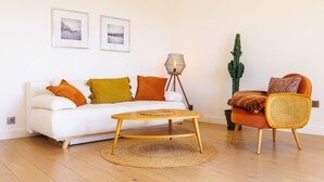 A living room with a white sofa, orange and yellow cushions, a round wooden coffee table, a wicker chair, a cactus, and two framed pictures on the wall. - Apt le NID - Sea view in the village (Tourrettes-sur-Loup)