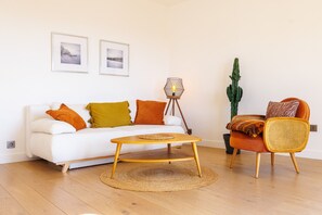 A living room with a white sofa, orange and yellow cushions, a round wooden coffee table, a wicker chair, a cactus, and two framed pictures on the wall. - Apt le NID - Sea view in the village (Tourrettes-sur-Loup)