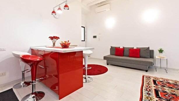 A modern kitchen with a red island and bar stools, a grey sofa with red cushions, and a patterned rug. - SUITE BOSCHETTO HOLIDAYS, warm apartment in the historic center of Rome (Roma)