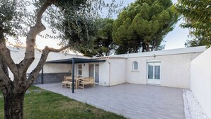 Une terrasse couverte avec une table et des chaises en bois, un bâtiment blanc avec une porte, et un arbre au premier plan.