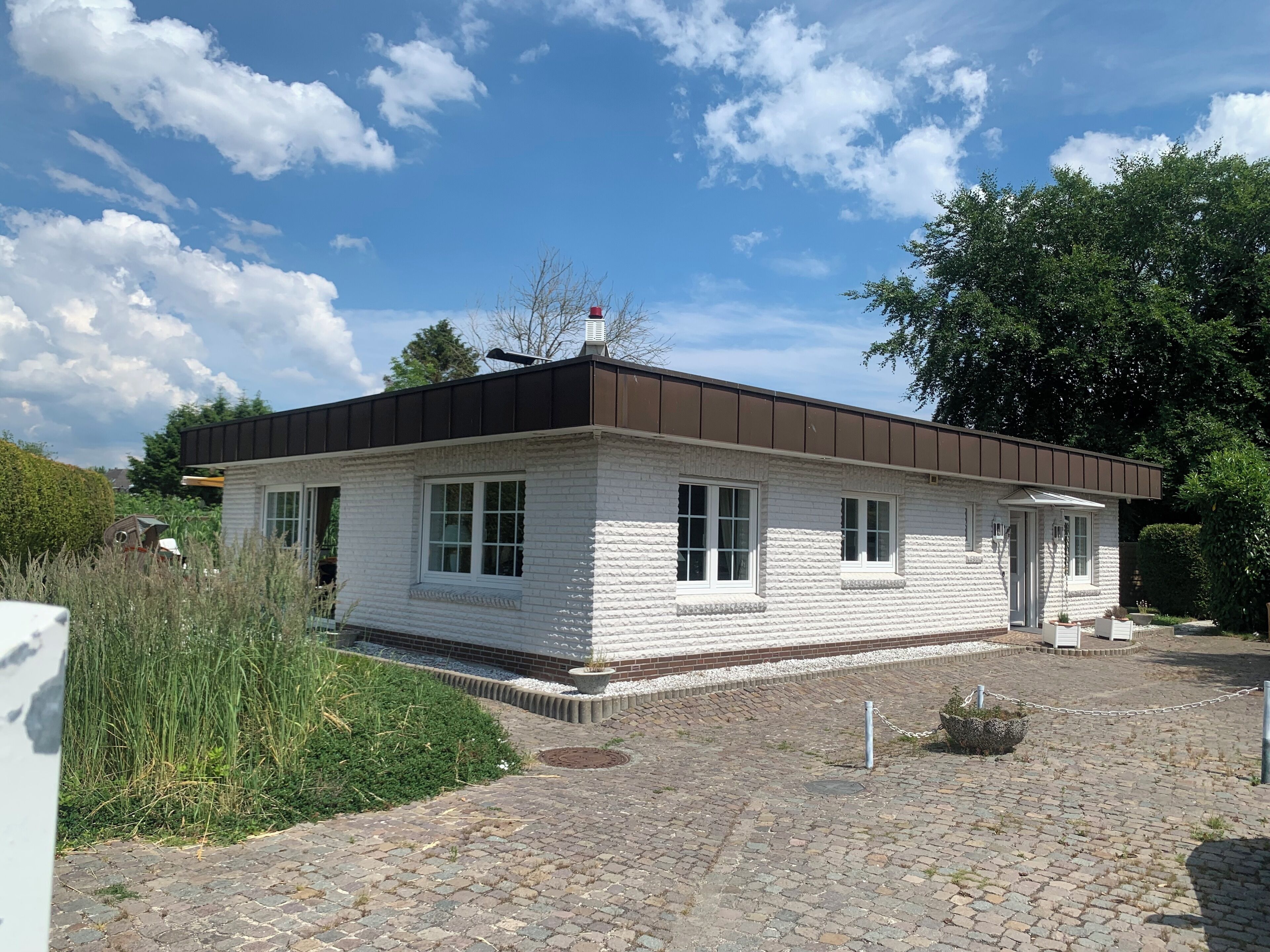 Ein eingeschossiges, weißes Gebäude mit einem braunen Dach, umgeben von einer gepflasterten Fläche und Grünflächen.