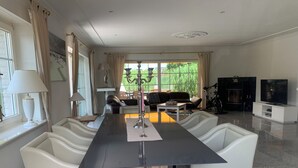 A spacious living room with a long dining table, white leather chairs, a sofa, and a TV. - Spacious vacation bungalow in Scharbeutz (125sqm) (Scharbeutz)