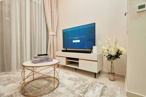 Una sala moderna con una televisión de pantalla plana sobre un mueble blanco, una mesa redonda con un libro y un jarrón con flores.