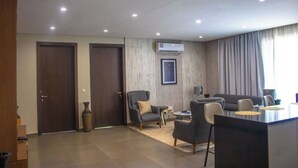 Ein modernes Wohnzimmer mit einer Couch, Sesseln, einem Couchtisch und einem kleinen Beistelltisch. Es gibt zwei Türen, ein großes Fenster mit Vorhängen und eine Klimaanlage an der Wand.