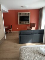 Habitación de hotel con cama, sofá, televisor montado en la pared, escritorio con silla y ventana con cortinas.