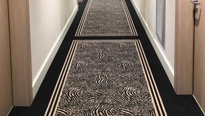 Un couloir d’hôtel avec un tapis à motifs noir et blanc, des portes en bois et des tableaux encadrés accrochés aux murs.