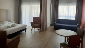 Une chambre d’hôtel avec un lit, une chaise, un canapé, une table ronde et de grandes fenêtres avec des rideaux.