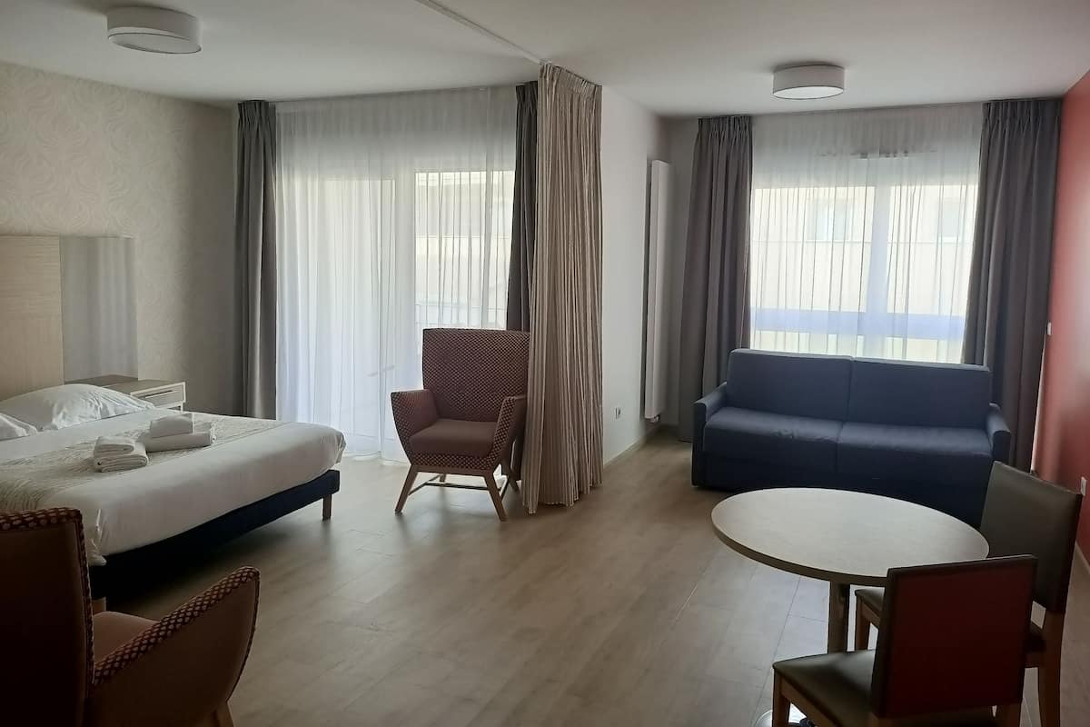 Una camera d'albergo con un letto, una sedia, un divano, un tavolo rotondo e ampie finestre con tende.