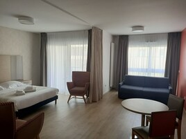 Habitación de hotel con cama, sillón, sofá, mesa redonda y ventanas grandes con cortinas.