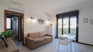 Ein Wohnzimmer mit einem beigen Sofa, einem weißen Tisch und Stühlen. Ein Fenster bietet Blick auf Grünflächen.