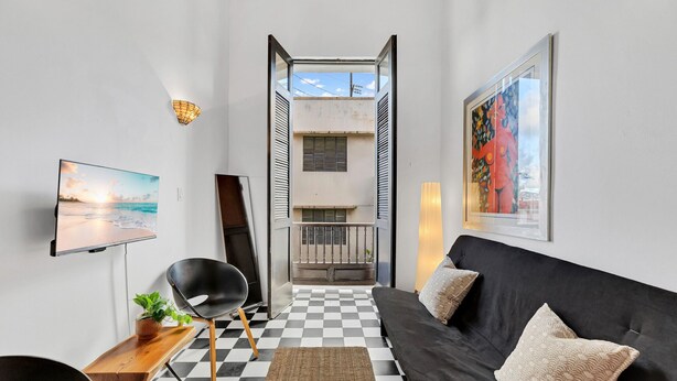 Un salon moderne avec un sol à carreaux noir et blanc, un canapé noir agrémenté de coussins à motifs, une table d’appoint en bois avec une plante, une chaise noire, un téléviseur fixé au mur et un tableau encadré.