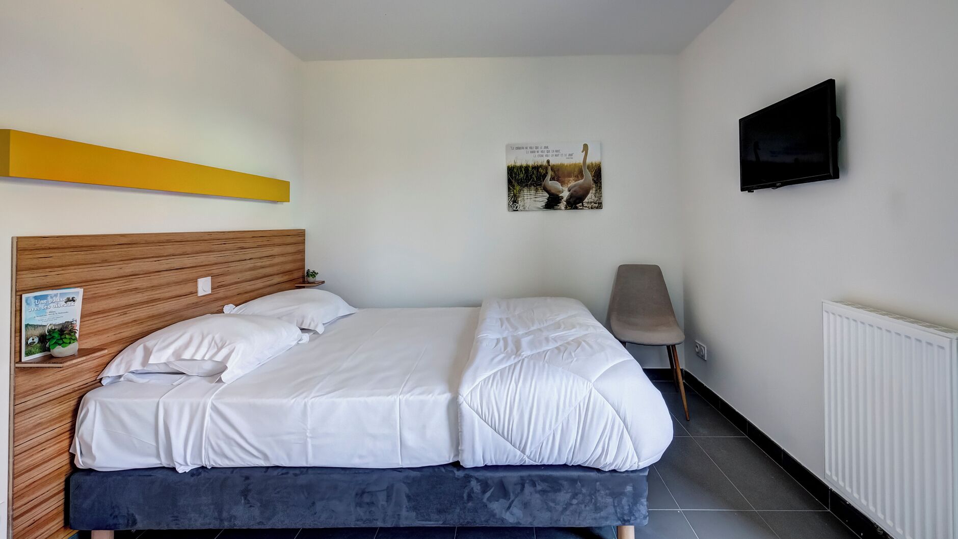 Une chambre avec une tête de lit en bois, un lit avec une literie blanche, une petite table de chevet avec une plante, une affiche accrochée au mur et un téléviseur fixé au mur.