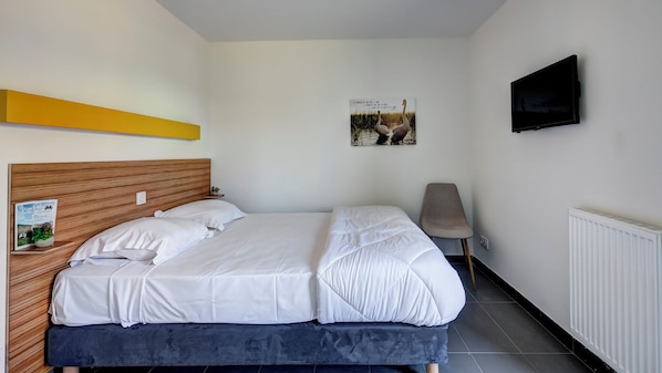A bedroom with a wooden headboard, a bed with white bedding, a small bedside table with a plant, a poster on the wall, and a TV mounted on the wall. - Vacancéole Les Chambres des Oiseaux (Motz)