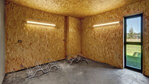 A room with wooden walls, a concrete floor, and metal chairs. There are two fluorescent lights on the ceiling, and a large window on the right side. - Vacancéole Les Chambres des Oiseaux (Motz)