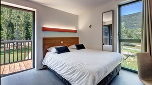 A modern bedroom with a large bed, a wooden headboard, and a view of a forest through a sliding glass door. - Vacancéole Les Chambres des Oiseaux (Motz)