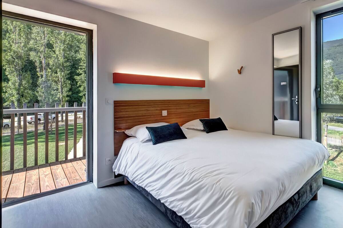 A modern bedroom with a large bed, a wooden headboard, and a view of a forest through a sliding glass door.