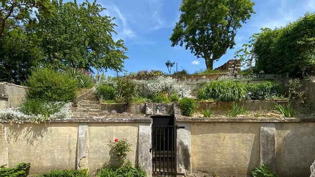 Cortile lastricato con un cancello, circondato da vegetazione e un muro in pietra.