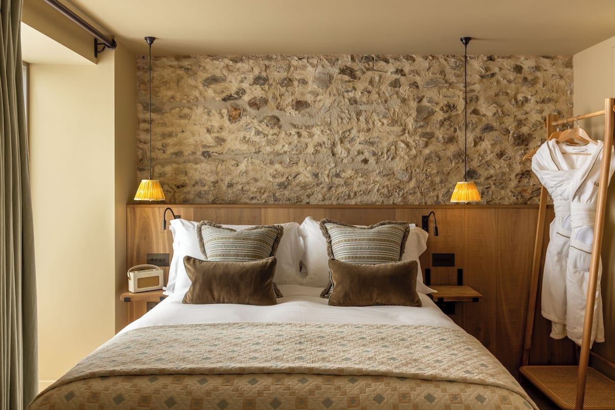 Ein Hotelzimmer mit Steinwand, einem Bett mit gemusterten Betttextilien, zwei Nachttischlampen, einem Holznachttisch mit Radio und einem Bademantelständer.