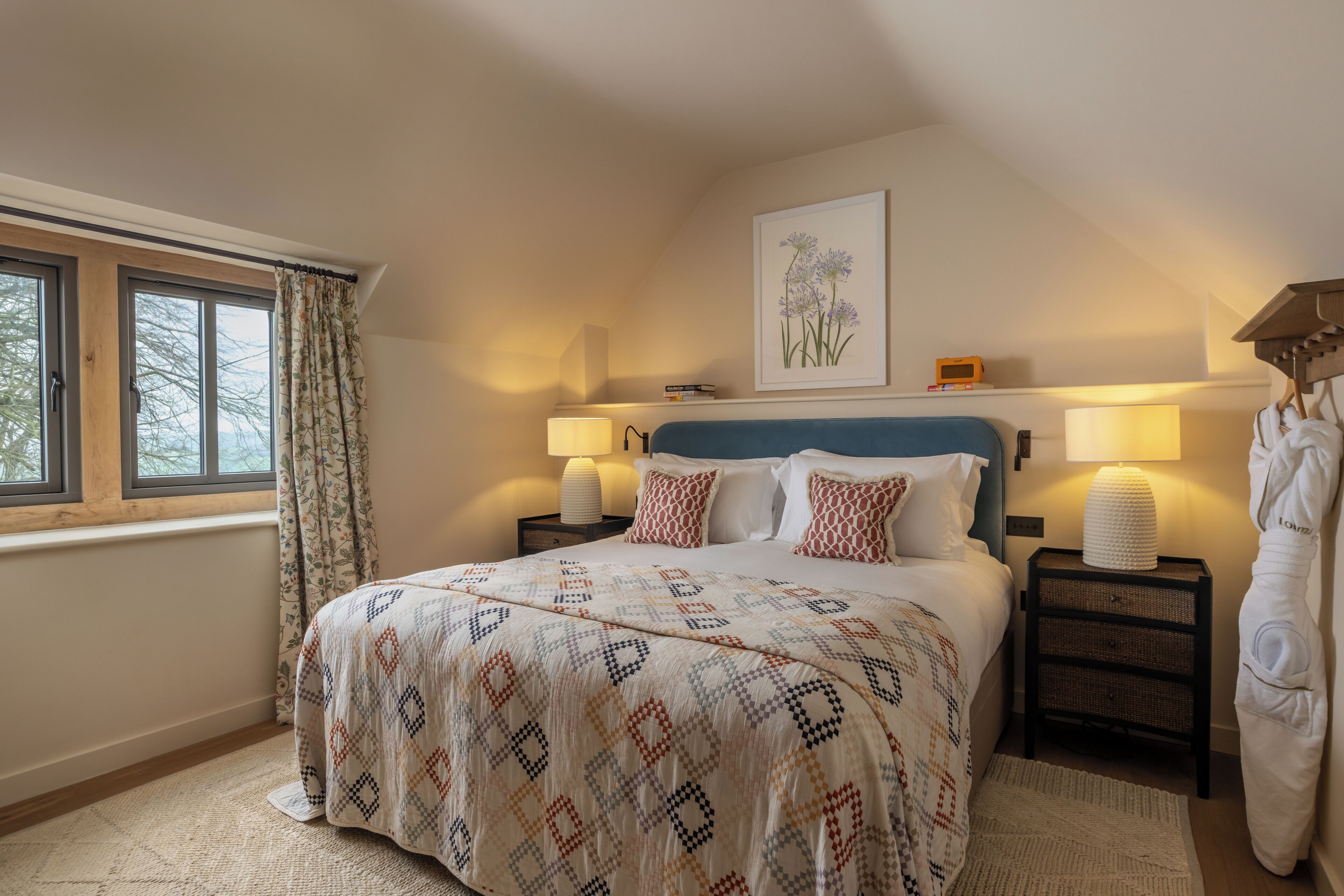 A bedroom with a bed, two bedside tables, a window with curtains, a wall-mounted lamp, and a framed picture.