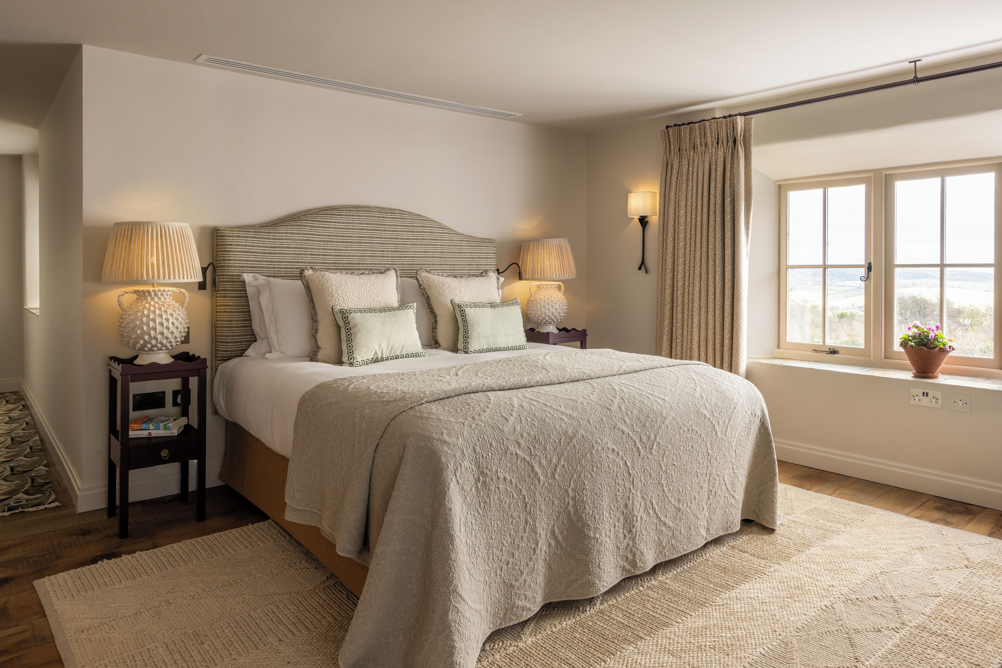 A bedroom with a large bed, two bedside tables with lamps, a window with curtains, and a view of the outdoors.