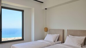Une chambre avec deux lits, une grande fenêtre donnant sur la mer et un luminaire suspendu.