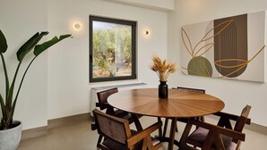 Un espace repas moderne avec une table ronde en bois, six chaises, une plante en pot, un vase avec des fleurs séchées, une grande fenêtre donnant sur un paysage verdoyant et une œuvre d’art murale.
