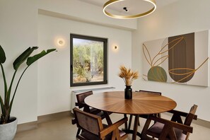 Un espace repas moderne avec une table ronde en bois, six chaises, une plante en pot, un vase avec des fleurs séchées, une grande fenêtre donnant sur un paysage verdoyant et une œuvre d’art murale.