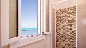Ein Badezimmer mit Fenster zum Strandblick, einer Dusche mit einer Wand aus Holzlatten und einem gefliesten Boden.