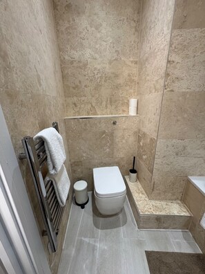 A bathroom with a toilet, a towel rack, a toilet brush, and a shelf with a roll of toilet paper. - Clockwork Stay (London)