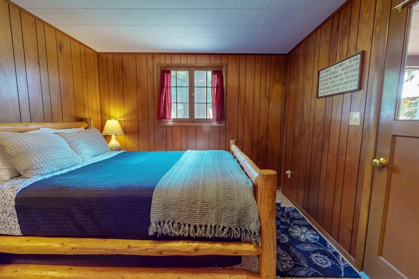 A wooden-paneled room with a bed, a window with curtains, and a door with a sign.