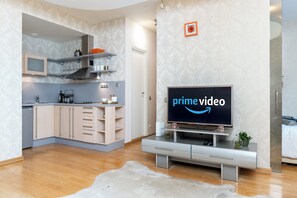 Moderni olohuone, jossa on litteänäyttöinen televisio, jonka näytöll ä näkyy Prime Videon logo, vaaleanvärisillä kaapistoilla varustettu keittiö ja sisäänrakennettu uuni sekä suuri valkoinen matto puulattialla.