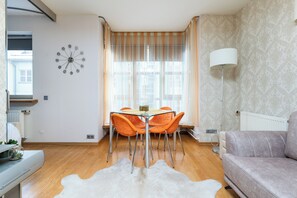 Moderni olohuone, jossa on ruokapöytä, oranssit tuolit, valkoinen sohva, tekoturkismatto, kello ja lattiavalaisin.