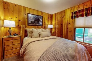 A wooden-paneled bedroom with a bed, two bedside tables, lamps, a framed picture, and a window with blinds.