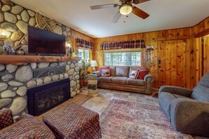 A cozy living room with a stone fireplace, wooden walls, a television mounted above it, and a ceiling fan.