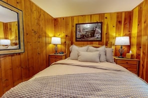 A wooden-paneled room with a bed, two bedside tables with lamps, a framed picture on the wall, and a mirror on the left wall.