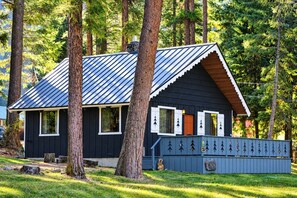 A cabin with a metal roof, surrounded by trees, and a fenced area with a bench and a small statue.