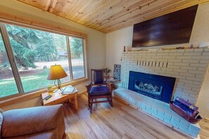 A cozy room with a fireplace, a television mounted above it, a window with a view of greenery, and a small table with a lamp. - Frazier Lake | Wallowa Lake rental (Joseph)