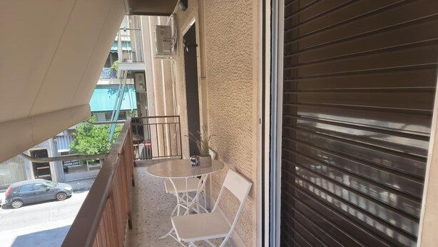 A balcony with a table and chairs, a view of the street, and a closed shutter door. - Casa Dili Stylish 2BR Apartment (Athens)