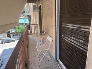 A balcony with a table and chairs, a view of the street, and a closed shutter door. - Casa Dili Stylish 2BR Apartment (Athens)