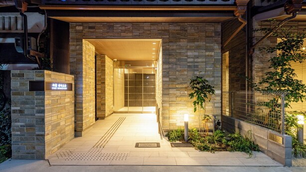A modern hotel entrance with a stone facade, a covered entrance, and a small garden area with a fence and plants. - oyado shinmachi gojo (Kyoto)