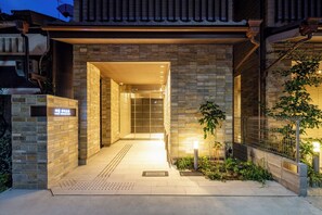 Una entrada de hotel moderna con fachada de piedra, un acceso cubierto y un pequeño jardín con cerca y plantas.