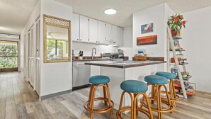 A modern kitchen with white cabinets, a central island with stools, a large mirror, and a potted plant. - *Kalanikai 215 | Kona Condo Near Downtown, Beaches and Dining (Kailua Kona)