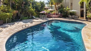 A swimming pool with a stone deck, surrounded by lush greenery and outdoor seating areas. - *Kalanikai 215 | Kona Condo Near Downtown, Beaches and Dining (Kailua Kona)