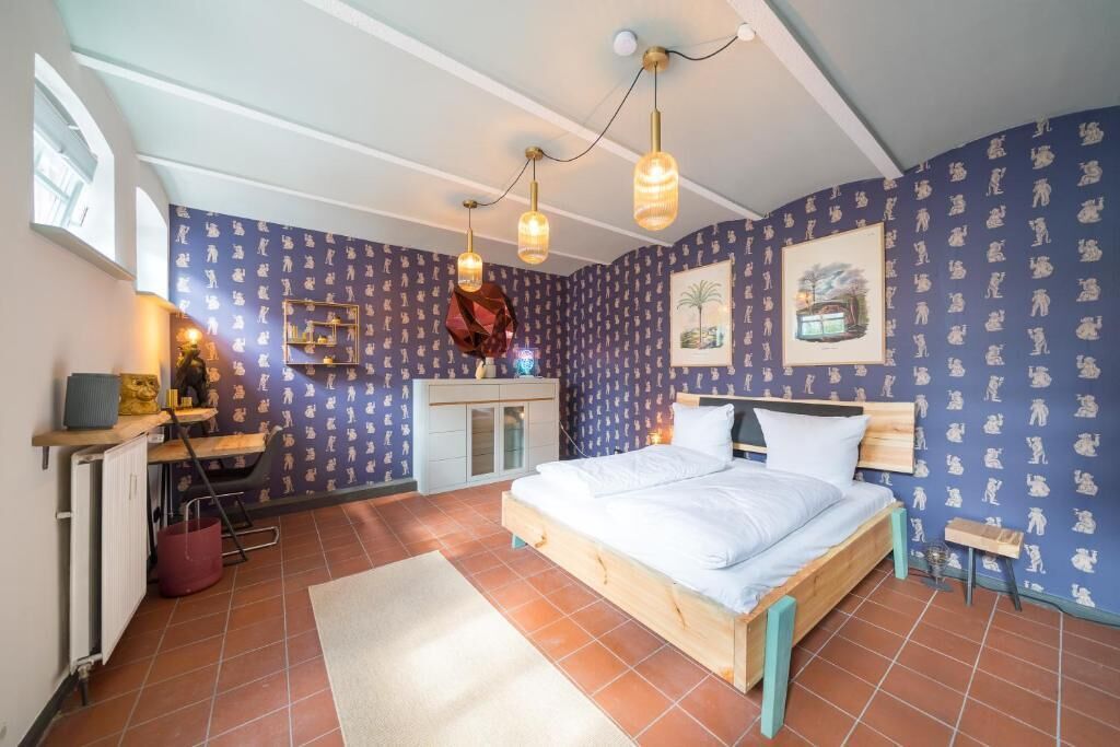 A bedroom with a bed, a desk, and a chair. There are two wall-mounted lights and a window.